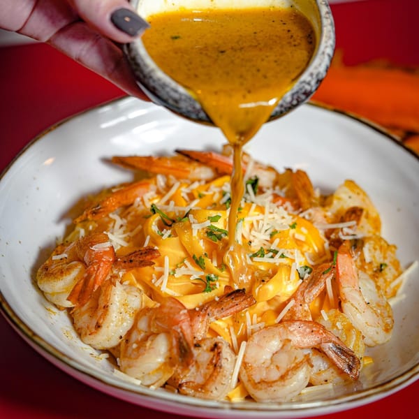 Peruvian shrimp pasta with a creamy yellow sauce being poured over it, garnished with cheese and herbs.