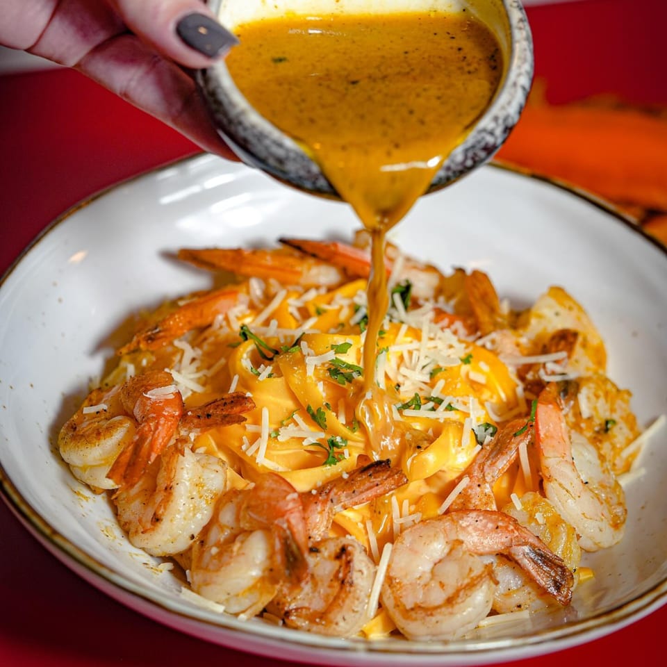 Peruvian shrimp pasta with a creamy yellow sauce being poured over it, garnished with cheese and herbs.