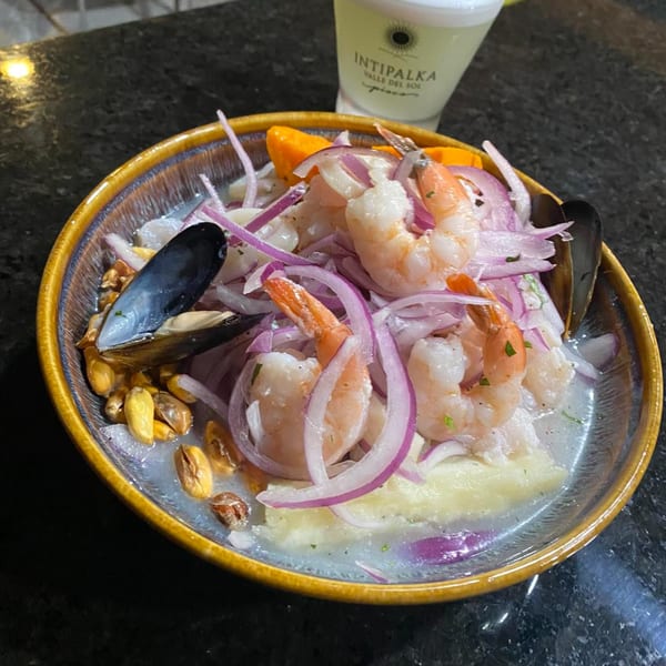 Peruvian ceviche with shrimp, mussels, red onion, and corn in a bowl, with a drink in the background.