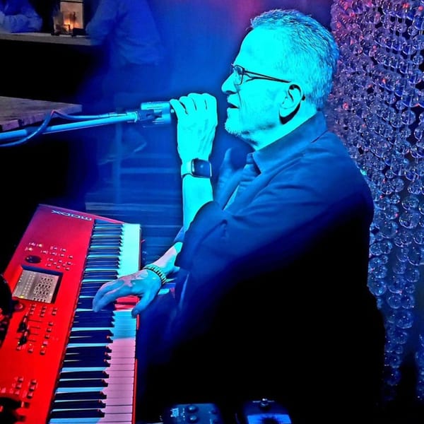 Musician singing into a microphone while playing a red keyboard, illuminated by blue stage lights.
