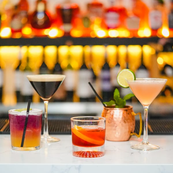 Four colorful cocktails and a copper mug with mint on a white marble bar.