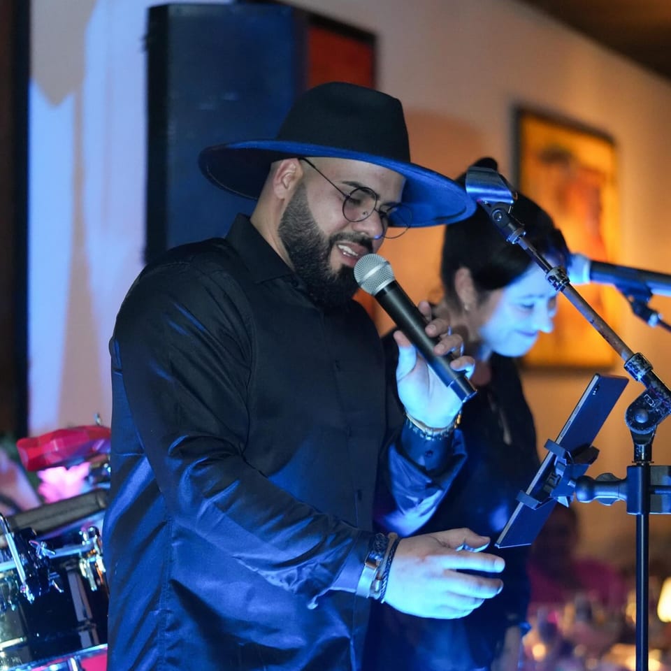 A male singer in a black hat and glasses performs with a microphone under blue stage lighting.