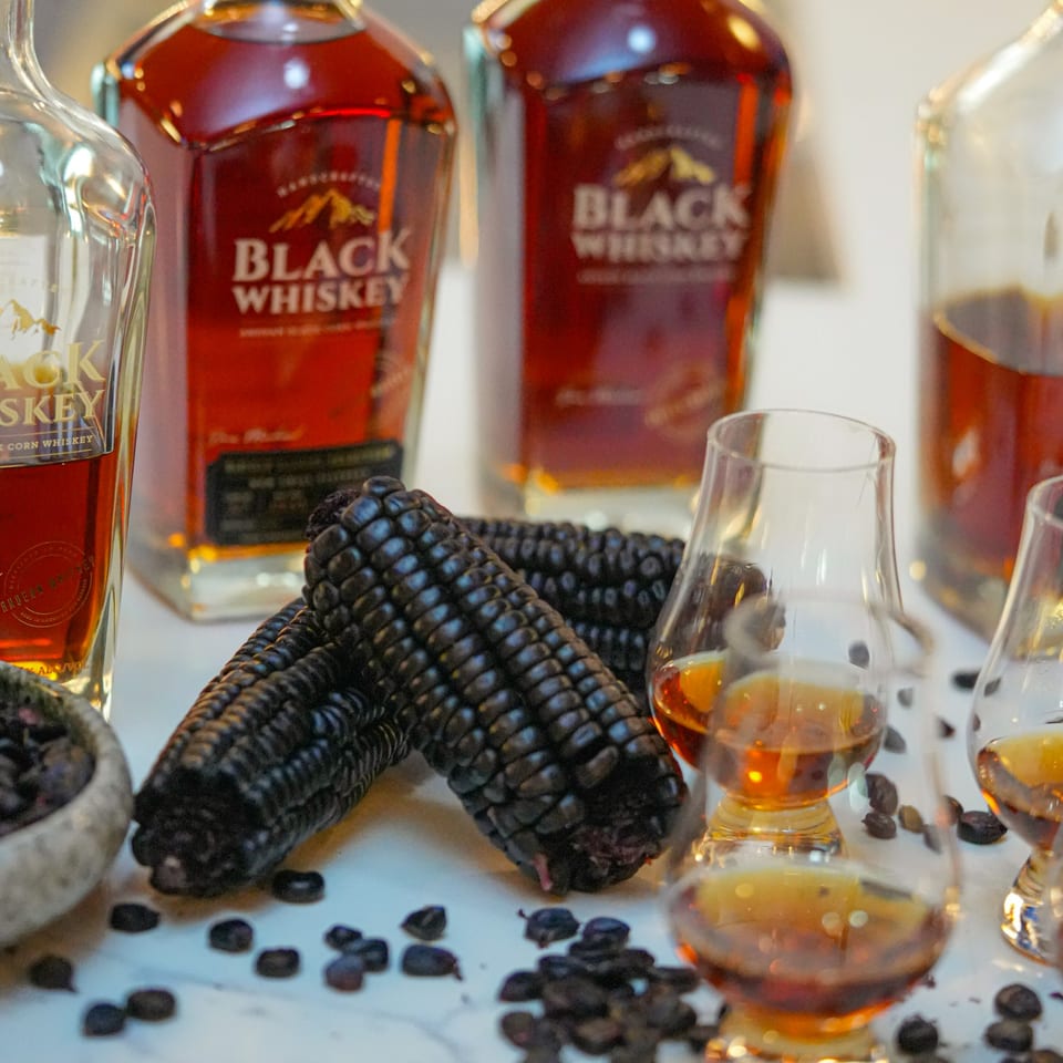 Bottles of "Black Whiskey" and glasses of whiskey with purple corn on a white surface.