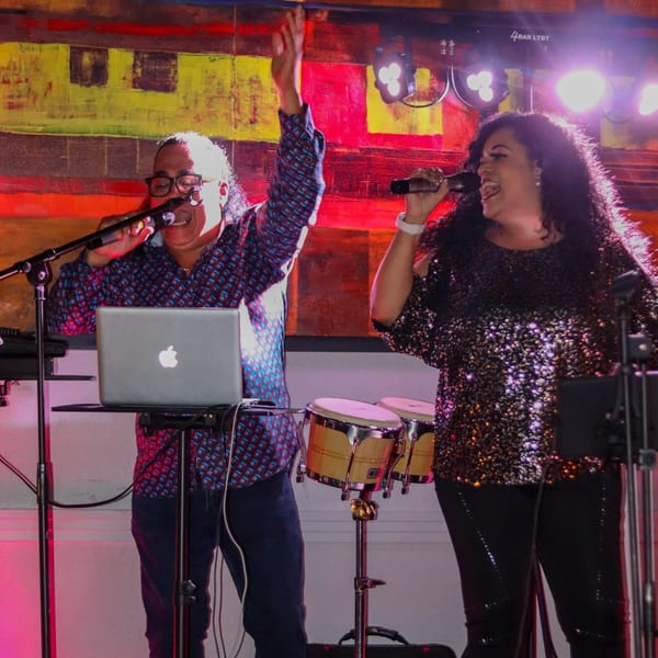 Two singers, a man and a woman, perform live on stage with a laptop and bongos.