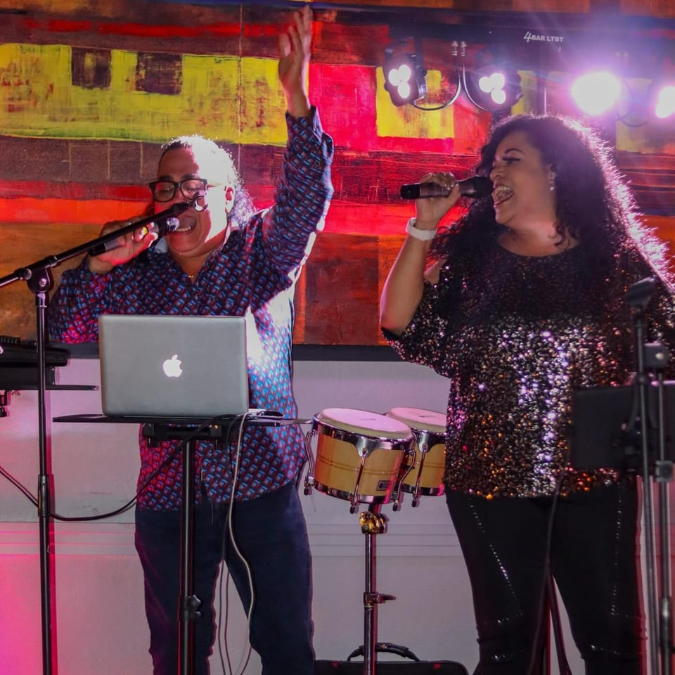 Two singers, a man and a woman, perform live on stage with a laptop and bongos.