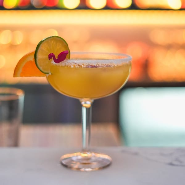 A Peruvian cocktail, garnished with lime, orange, and a flamingo pick, sits on a bar.