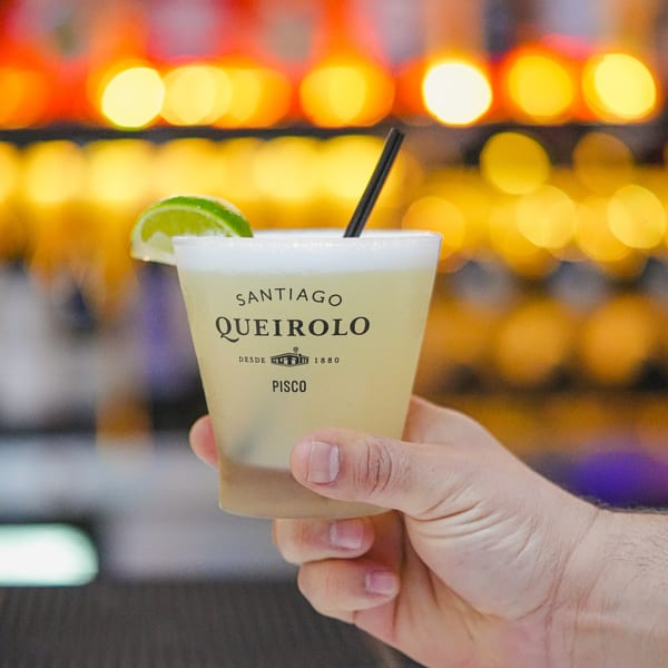 Hand holding a Santiago Queirolo Pisco cocktail with a lime wedge and straw, against blurred lights.