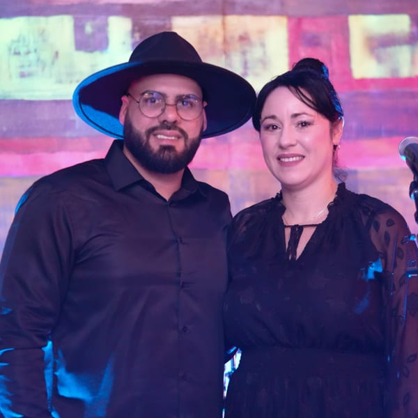 A man in a hat and a woman smiling, standing together in front of a colorful, abstract background.