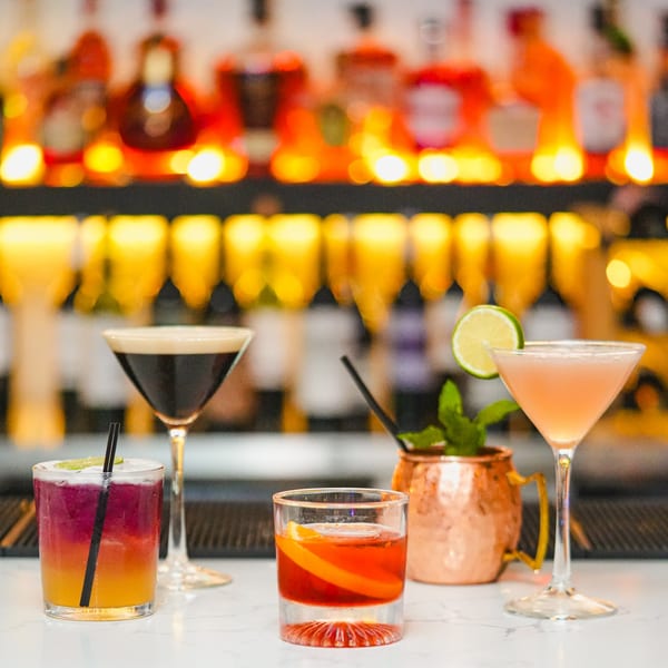 Five colorful cocktails, including a Pisco Sour, on a bar with blurred liquor bottles in the background.