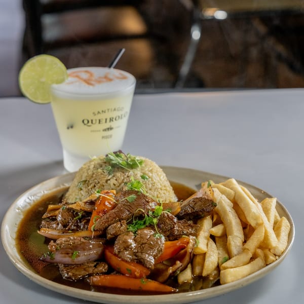Peruvian Lomo Saltado with steak, fries, rice, and a Pisco Sour cocktail with a lime wedge.