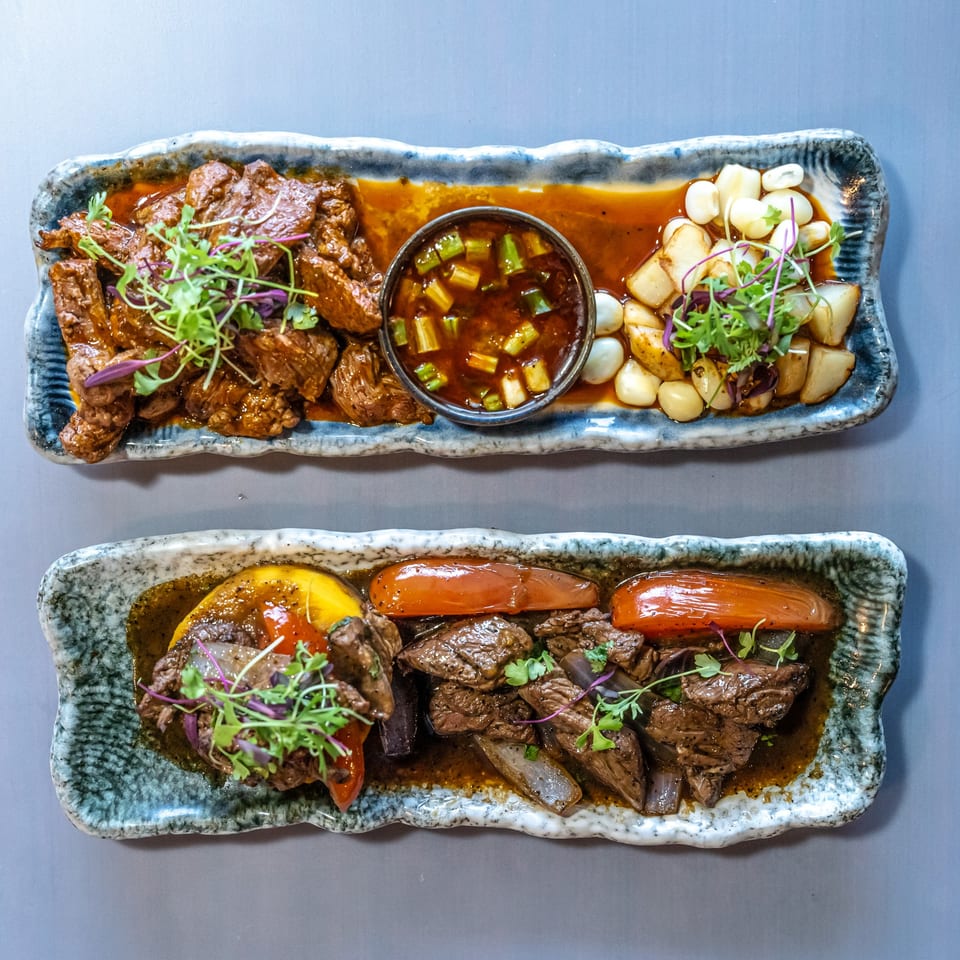 Two rectangular plates of Peruvian beef stew, one with corn and potatoes, the other with peppers.