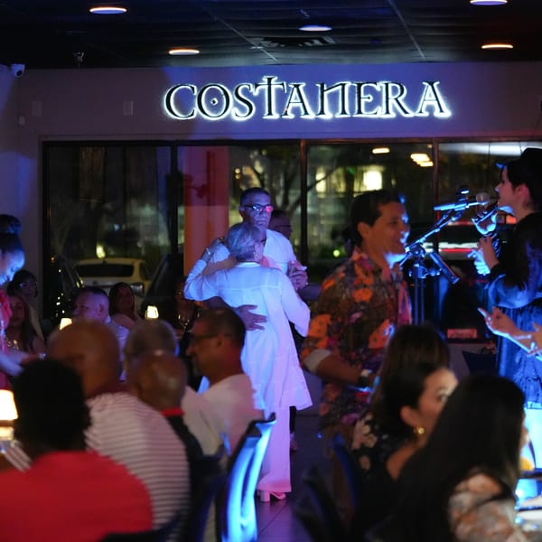 Couples dancing and a band performing under the glowing "Costanera" sign at night.