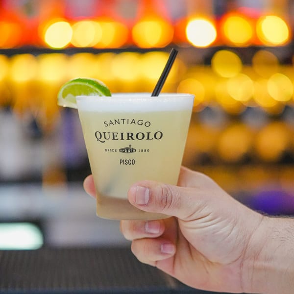 Hand holding a Pisco Sour cocktail with a lime wedge and black straw, against blurred lights.